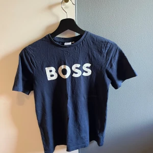 Mörkblå BOSS t-shirt XS - Snygg mörkblå t-shirt från BOSS i storlek XS med vit BOSS-logga på bröstet. Klassisk rund hals och korta ärmar. Tillverkad i mjuk bomull som känns skön mot huden. Perfekt för dig som gillar stilrena och enkla plagg med tydlig branding.