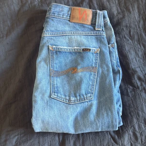 Bootcut jeans från Nudie, ljusblå - Säljer ett par ljusblå bootcut jeans från Nudie Jeans i modellen Bootcut Barry. Jeansen är i 100% bomull med klassisk femficksdesign, orange sömmar och snygg slitning. Märkespatch bak och vågig brodyr på bakfickan. Perfekta för dig som gillar retrovibbar.