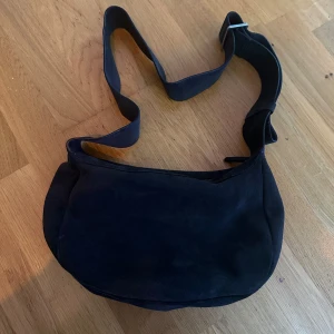 Svart axelväska i mocka från COS - Säljer en stilren svart axelväska från COS i mjuk mocka. Väskan har justerbar rem, dragkedja upptill och rymligt fack med innerficka. Perfekt för dig som gillar minimalistisk design och vill ha en väska som rymmer allt du behöver.