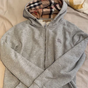 Grå hoodie från Burberry med rutig huva - Snygg grå hoodie från Burberry med dragkedja och klassisk rutig insida i huvan. Tröjan har långa ärmar, huva med snörning och ett litet Burberry-logo broderat på bröstet. Perfekt för dig som gillar stilrena och lyxiga detaljer.