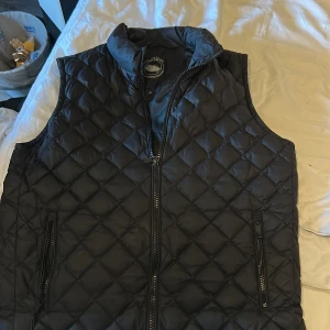 Svart quiltad dunväst från Saki Sweden - Svart dunväst från Saki Sweden med quiltat mönster och hög krage. Västen har två fickor med dragkedja och hel dragkedja framtill. Perfekt för lager-på-lager och ger en clean, stilren look nypris 1299 sitter som M.