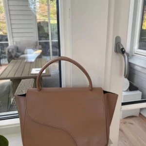 Beige handväska i skinn med axelrem - Snygg ATP atelier väska i modellen arezzo med både handtag och avtagbar axelrem. Väskan har en snygg asymmetrisk lockdesign och guldiga metalldetaljer. Perfekt storlek för det viktigaste och ger en clean look till din outfit. Nypris 5200!