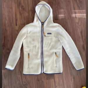 Vit teddyjacka från Patagonia - Säljer en vit teddyjacka från Patagonia med huva och blå detaljer. Jackan har dragkedja framtill, tre fickor med dragkedja och mysigt fleece-material. Perfekt för kyliga dagar och har en clean, sportig look.