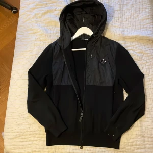 Svart hybrid jacka från J.Lindeberg - Svart vindjacka från J.Lindeberg med huva och hel dragkedja. De är en mestadels stickad jacka med polyester där uppe. Passar perfekt hela året. Finns lite färgstänk längst ner på höger ärm men inget man märker. NYPRIS 3000 köpt i butik Stockholm 