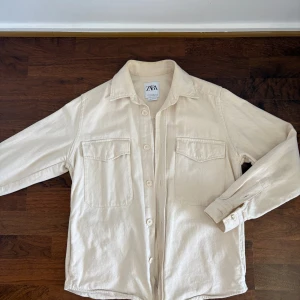 Beige overshirt från Zara med print - Snygg beige overshirt från Zara i bomull med relaxed fit. Två stora bröstfickor framtill och knäppning med knappar. På ryggen finns ett coolt print med palmer i brunt och svart. Perfekt för lager-på-lager och chill stil samt hösten och våren. 