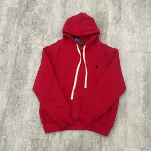 Röd hoodie från Polo Ralph Lauren - Riktigt snygg röd hoodie från Polo Ralph Lauren med klassisk broderad logga på bröstet. Tröjan har vit snörning i huvan, ribbade muddar och är tillverkad i mjuk bomull. Perfekt för dig som vill ha en stilren och bekväm look.