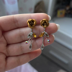 Guldros örhängen med kristaller - Unika örhängen med guldfärgade rosor upptill och silverfärgade stjälkar dekorerade med glittrande gula och klara kristaller. Blad och detaljer i silver ger en elegant och kreativ look. Perfekt för dig som vill sticka ut med ett blommigt smycke.