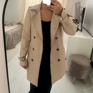 trenchcoat  - Stilren beige trenchcoat från Zara Woman med dubbelknäppning, klassisk krage och bälte i midjan samt vid ärmslut. Jackan har snygga detaljer som axelklaffar och fickor framtill. Perfekt för dig som vill ha en tidlös och trendig look.
