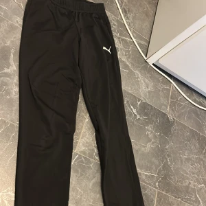 Svarta träningsbyxor från Puma - Svarta träningsbyxor från Puma med elastisk midja och diskret Puma-logga på låret. Byxorna är tillverkade i ett lätt och smidigt syntetmaterial som passar perfekt för sport och aktivitet. Klassisk rak passform och stilren look.