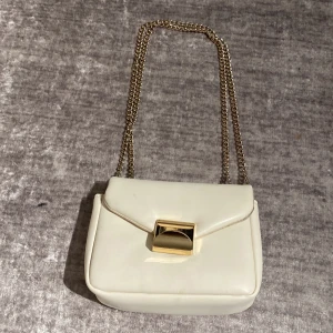 Vit axelväska med guldkedja - Stilren vit axelväska med kedja i guld och snyggt guldfärgat spänne framtill. Väskan har en klassisk, boxig form och är tillverkad i ett slätt material som ger en lyxig känsla. Perfekt för dig som vill ha en clean och trendig look.