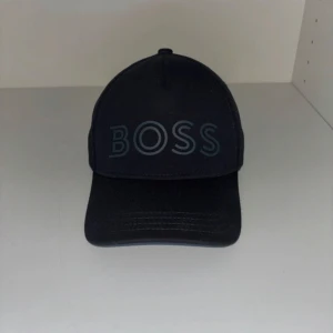 Svart keps från BOSS - Snygg svart keps från BOSS med böjd skärm och stort BOSS-tryck framtill i grått. Klassisk form med paneler och synliga sömmar. Perfekt för dig som vill ha en stilren och modern look. 