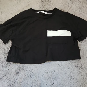 Svart croppad t-shirt Calvin Klein - Svart croppad t-shirt från Calvin Klein Jeans med vit logga över bröstfickan. T-shirten har korta ärmar, rund halsringning och är gjord i mjuk bomull. Perfekt för en chill och trendig look.