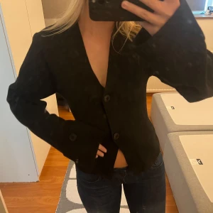 Svart cardigan från Gina Tricot - Snygg svart cardigan från Gina Tricot med v-ringad hals och stora knappar framtill. Jackan har lång ärm och är tillverkad av 51% återvunnen polyester. Stilren och enkel modell som är lätt att matcha med jeans eller kjol. Helt ny aldrig använd