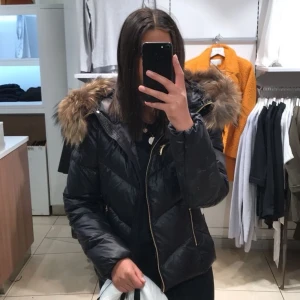 Hollies puffer jacka  - Snygg svart pufferjacka med glansig finish och stor huva med fluffig päls. Jackan har guldiga dragkedjor. Perfekt för kalla dagar när du vill vara både varm och stilren, Pris kan diskuteras 