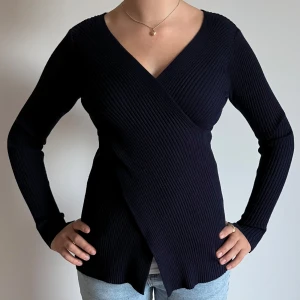 Mörkblå omlottstickad tröja - Säljer en mörkblå ribbstickad omlott-tröja med lång ärm och v-ringad hals. Tröjan har en snygg omlott-design som ger en schysst siluett och är tillverkad i ett stretchigt stickat matrial.