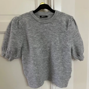 Grå stickad tröja med puffärm Gina Tricot - Grå stickad tröja från Gina Tricot med korta puffärmar och rund halsringning. Tröjan har en mjuk och mysig känsla, ribbade muddar och en klassisk, enkel design som passar perfekt till jeans eller kjol.