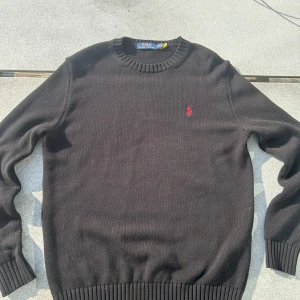 Ralph Lauren Stickad Tröja - Svart stickad tröja från Polo Ralph Lauren med klassisk rund halsringning och röd broderad logga på bröstet. Långärmad modell med ribbade muddar och nederkant. Perfekt för dig som gillar stilrena och tidlösa plagg.