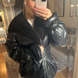 Svart pufferjacka med pälsdetalj - Säljer en svart pufferjacka med glansig yta och bred krage i mjuk pälsimitation. Jackan har oversized passform, stora puffiga ärmar och knappar framtill. Snygga detaljer med spännen vid ärmsluten och markerad midja. Perfekt för dig som gillar statement-plagg.