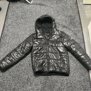 Svart pufferjacka från Jordan - Säljer en svart pufferjacka från Jordan med ikoniska Jumpman-loggan på bröstet och ärmen. Jackan har huva, dragkedja framtill och är tillverkad i glansig polyester med mjukt foder. Perfekt för kalla dagar när du vill ha en sportig och clean look. Priset kan också diskuteras