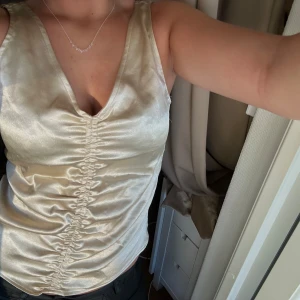 Champagnefärgad snygg topp - Passar mig som är XS/S. Snygg champagnefärgad linnetopp med omlott-effekt och v-ringning. Toppen har en glansig satinlook och är ärmlös med snygg rynkning längs mitten framtill. Perfekt för dig som vill ha en trendig och chic look.