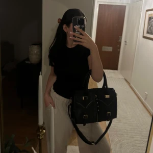 Svart handväska med gulddetaljer - Snygg svart handväska i skinnimitation med både handtag och axelrem. Väskan har flera guldfärgade detaljer som spännen, dragkedjor och dekorativa nitar framtill. Stilren och rymlig modell med två ytterfickor och en klassisk boxig form.