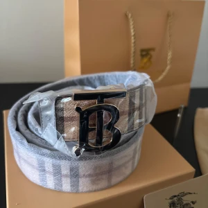 Rutig Burberry bälte med silverdetalj - Snyggt bälte från Burberry med klassiskt rutigt mönster i beige, grått och rött. Bältet har en stor metallspänne med Burberrys ikoniska TB-logga i silver. Perfekt accessoar för att lyfta din outfit med en lyxig touch.