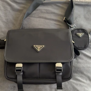 Svart axelväska  - Snygg svart axelväska från  med klassisk trekantig logga i metall på framsidan. Väskan har justerbar axelrem och ett extra litet fack med dragkedja som sitter fast på remmen. Tillverkad i slitstark nylon med detaljer i skinn och metallspännen.