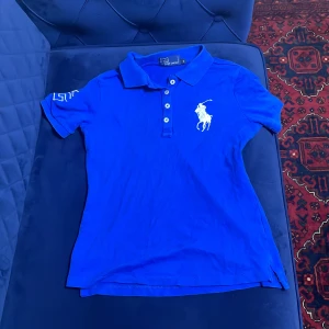 Blå Polo Ralph Lauren pikétröja S - Klassisk blå pikétröja från Polo Ralph Lauren med vit broderad logga på bröstet och siffra 2 på ärmen. Tröjan har krage, knappar framtill och är tillverkad i bomull. Perfekt för en sportig och stilren look.
