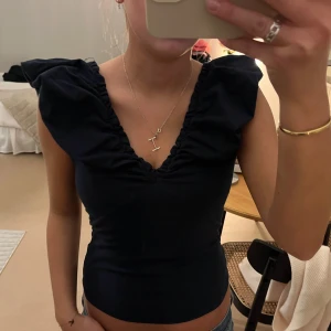 Svart volangtopp från Holly & Whyte - Supersnygg svart topp från Lindex med djup v-ringning och stora volangärmar. Toppen är figurnära och har en lätt rynkad detalj längs halsringningen. Storlek S, mycket bra skick!
