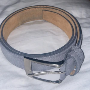 Grå mockabälte med silverfärgat spänne - Snyggt grått bälte i mocka med klassiskt silverfärgat metallspänne. Bältet har stilrena sömmar längs kanterna och insidan är brun. Perfekt accessoar för att lyfta din outfit.