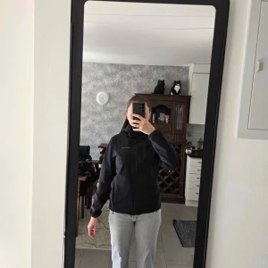 Svart vindjacka Peak Performance - Svart vindjacka från Peak Performance med huva och dragkedja framtill. Jackan har två sidofickor med dragkedja och justerbar snörning vid huvan. Stilren design och normal passform, perfekt för blåsiga dagar.