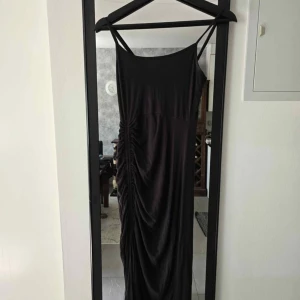 Svart långklänning med rynkade detaljer - Svart långklänning med smala axelband och snygga rynkade detaljer längs ena sidan. Klänningen har en enkel, elegant siluett och är perfekt för dig som gillar minimalistisk stil. Materialet känns mjukt och följsamt mot kroppen.