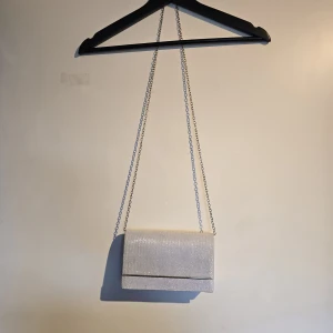 Glittrig silver kuvertväska med kedja - Snygg kuvertväska i glittrigt silver med lång kedjerem. Väskan har ett stilrent, rektangulärt format och stängs med magnetknapp. Insidan är ljus och har ett litet innerfack för kort eller småsaker. Perfekt för att lyfta din outfit med extra bling.