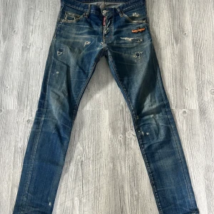 Dsquared2 blå slitna jeans med patch - Säljer ett par blå jeans från Dsquared2 med slitningar, hål och en cool kanot-patch vid fickan. Jeansen har klassisk femficksdesign, låg midja och smal passform. Materialet är denim i bomull och de har en snygg tvättad look med slitna detaljer. Väldigt slitna där av priset 
