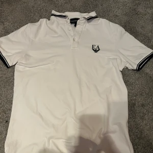 Vit piké från Emporio Armani - Vit pikétröja från Emporio Armani med svart EA-logga på bröstet. Klassisk krage med svarta ränder och knäppning framtill. Ärmarna har svarta detaljer vid mudden. Perfekt för en clean och stilren look.