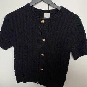 Svart kabelstickad kofta med guldknappar - Kortärmad svart kabelstickad kofta med rund hals och snygga guldknappar framtill. Tröjan har en klassisk ribbad kant vid hals och knappslå, perfekt för lager-på-lager och en stilren look.