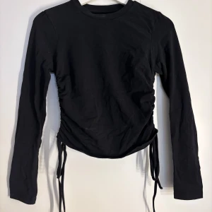 Svart långärmad topp med snörning - Svart långärmad topp i bomull med cool snörningsdetalj på sidorna som ger en snygg rynkad effekt. Från Pieces.