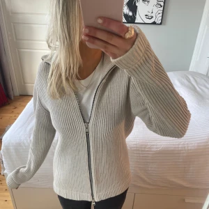 Beige stickad kofta med dragkedja - Ribbstickad beige kofta med hel dragkedja framtill och hög krage. Tröjan har lång ärm och är i ett mjukt, stickat material som känns både stilrent och bekvämt. Perfekt för lager-på-lager och passar till många olika outfits.