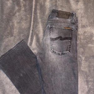 Nudie Jeans W27 L32 - Snygga grå jeans från Nudie Jeans Co med klassisk femficksdesign och svart broderad detalj på bakfickan. Jeansen har en rak passform och är tillverkade i bomull. Perfekta för dig som gillar en stilren och avslappnad look.