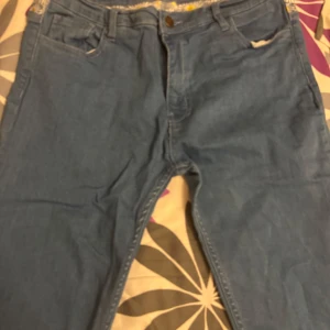 Blå jeans med broderade blommor BTW - Snygga blå jeans från BTW med raka ben och broderade vita och gula blommor längs linningen bak. Klassisk femficksmodell och läderpatch med logga. Perfekta för dig som vill ha något unikt och trendigt i garderoben.