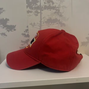 Röd keps från Dsquared2 - Riktigt snygg röd keps från Dsquared2 med broderad text 'No Mercy Dsquared2 Est. 1995' framtill i vitt, svart och gult. Klassisk böjd skärm och justerbar rem bak. Kepsen är tillverkad i bomull och har en sportig vibe. Priset ej hugget !