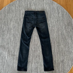 Mörkgrå jeans från Nudie Jeans, W29 L32 - Säljer ett par mörkgrå jeans från Nudie Jeans med klassisk femficksdesign och broderad bakficka. Jeansen har en rak passform och är tillverkade i mjukt bomullstyg. Perfekta för dig som gillar stilrena och tidlösa jeans.