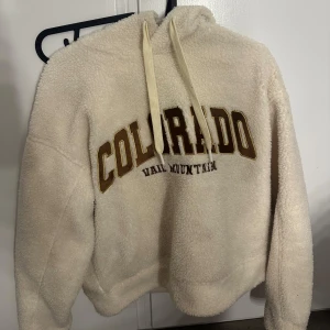 Beige teddy hoodie från Stay, S - Supermjuk och fluffig beige teddy hoodie från Stay med broderad text 'Colorado Vail Mountain' i brunt och gult framtill. Tröjan har huva med snörning och långa ärmar, perfekt för kyliga dagar. Oversized och cozy vibe.