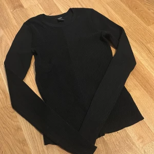 Svart ribbad tröja från Lindex - Svart långärmad tröja från Lindex i storlek S. Tröjan har en ribbad struktur på både fram- och baksida, rund halsringning och är figurnära i modellen. Perfekt att styla med jeans eller kjol för en clean look.