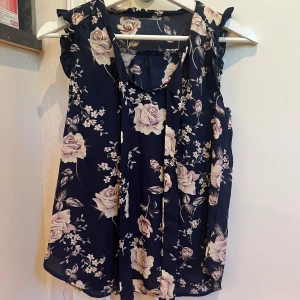 Blommig blus från Hallys i marinblått xs  - Supersöt blommig blus från Hallys i marinblått med ljusa rosor och småblommor. Ärmlös modell med volangdetaljer vid axlarna och knytband framtill. Luftigt material storlek xs 