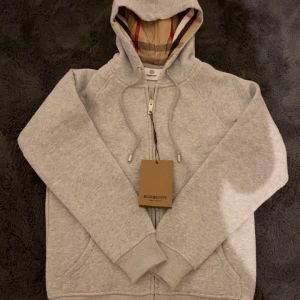 Grå huvtröja från Burberry - Säljer en grå hoodie från Burberry med klassiskt rutigt foder i huvan och broderad logga på bröstet. Tröjan har snörning i huvan, dragkedja framtill och ribbade muddar. Perfekt för dig som vill ha en stilren och lyxig look. ALDRIG använd och med original prislapp kvar. OBS: Liten i storleken, så tänk på det innan köp!
