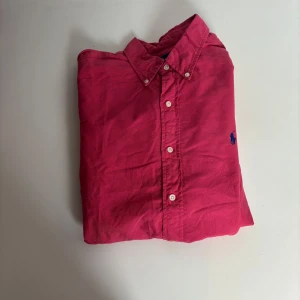 Rosa skjorta från Ralph Lauren L - Snygg rosa skjorta från Ralph Lauren i slim fit med klassisk krage och knäppning framtill. Skjortan har lång ärm och ikonisk blå broderad logga på bröstet. Tillverkad i mjuk bomull, perfekt för en clean och stilren look.
