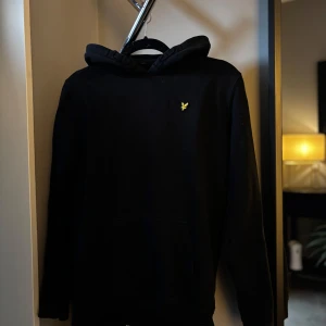 Svart hoodie från Lyle & Scott - Svart hoodie från Lyle & Scott med klassisk gul logga på bröstet. Tröjan har huva, känguruficka och långa ärmar. Perfekt för dig som gillar en clean och stilren look.