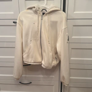 Vit hoodie från Moose Knuckles XS - Säljer en snygg vit hoodie från Moose Knuckles i storlek XS. Tröjan har dragkedja framtill, huva med snörning och en cool metallogo på ärmen. Mjuk insida och croppad passform gör den både bekväm och trendig. Använd några gånger 