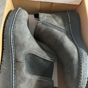 Grå chelsea boots , stl 39 - Snygga grå chelsea boots  med elastiska paneler på sidorna och dragkedja på insidan. Skorna har rund tå, platt sula och dekorativa vita sömmar längs sulkanten. Perfekta för dig som gillar stilrena och bekväma boots till vardags.
